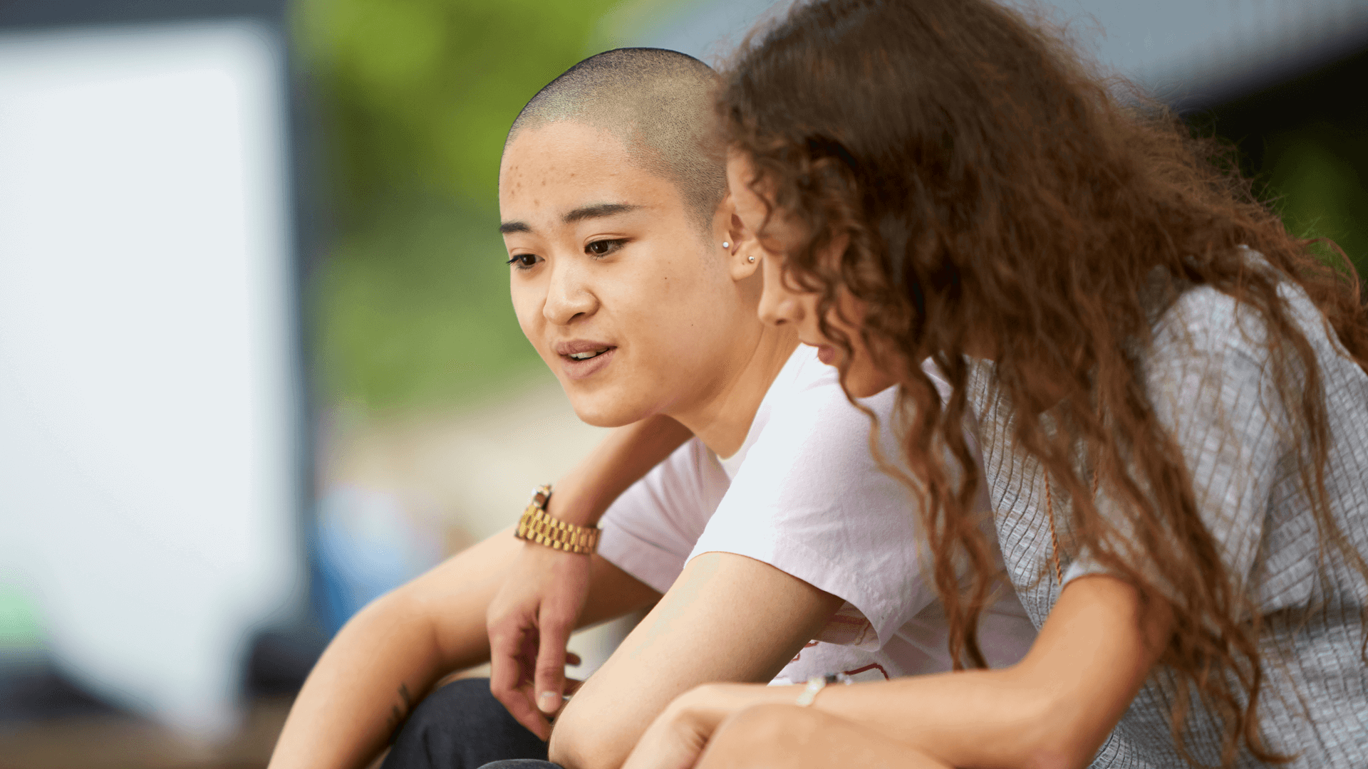 medium shot of a girl with curly hair wrapping her arms to her girlfriend talking to her while they are sitting in a park