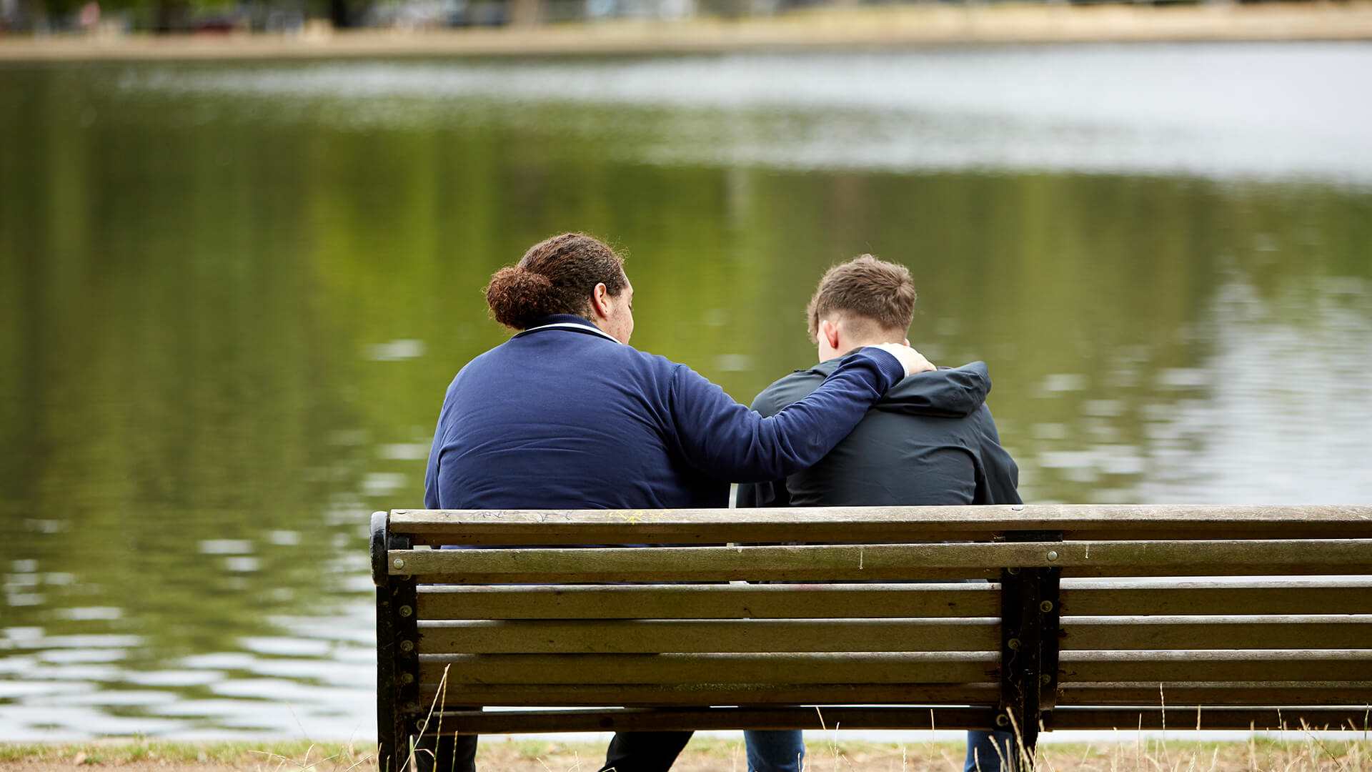 back-shot-of-two-boys-wearing-jackets-comforting-each-other-while-sitting-on-a-park-bench-in-front-of-a-lake