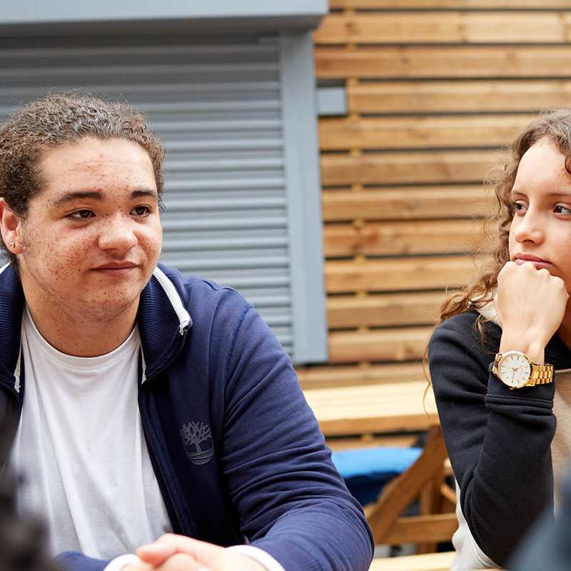 medium-shot-of-a-young-man-in-blue-jacket-and-white-shirt-looking-serious-while-sitting-with-a-girl-with-curly-hair-and-hands-on-her-chin-while-looking-at-him-and-sitting-on-a-bench-with-two-other-people-on-foreground