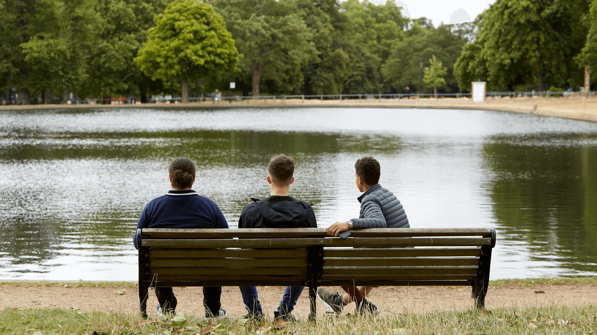 wide shot of three boys sitting on a bench looking over the lake in the park