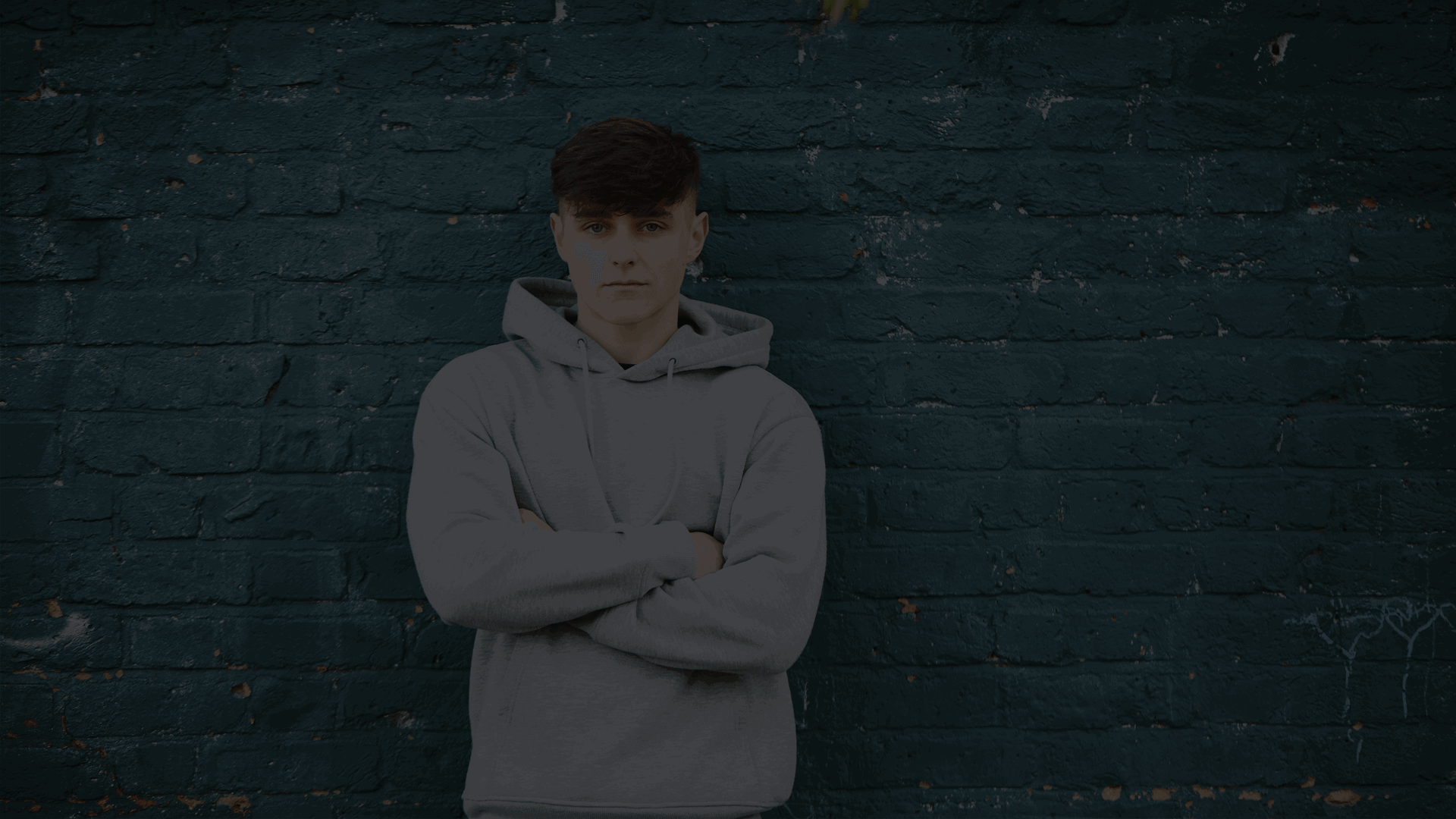 A young person stands with their arms folded leaning against a blue brick wall. They are looking serious at the camera.