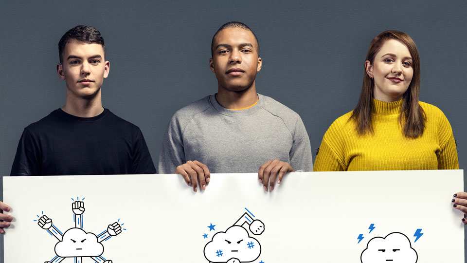 Three people hold up a large placard which has cartoon images of clouds on showing the emotions of outward aggression, inward aggression and passive aggression. The person on the left is wearing a black t-shirt looking at the camera with a neutral expression. The person in the middle is wearing a grey jumper looking neutral at the camera. The person on the right is wearing a yellow jumper and grinning at the camera.