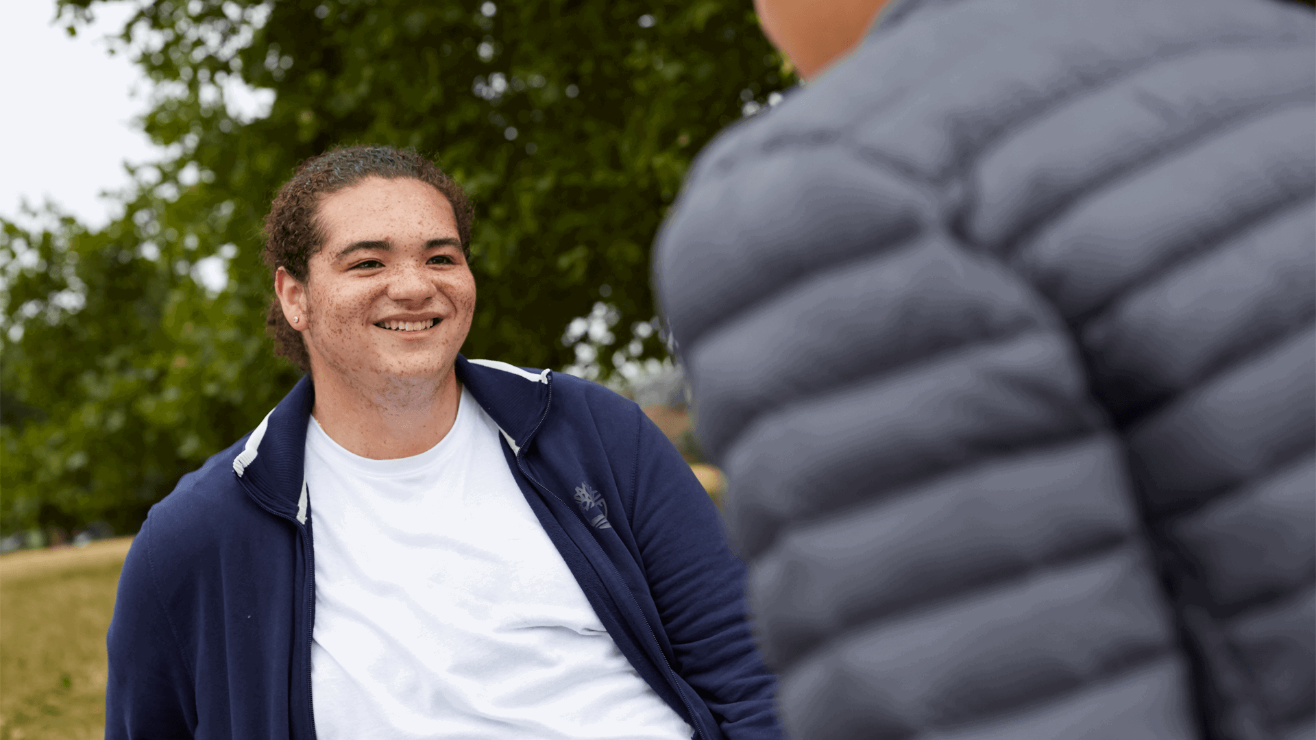 medium-shot-of-a-boy-wearing-blue-jacket-and-white-shirt-smiling-to-a-friend-with-trees-on-background