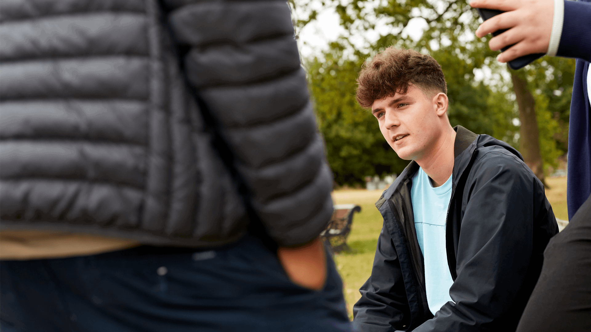 medium shot of a boy in black jacket and blue shirt sitting on bench in the park with people standing around him