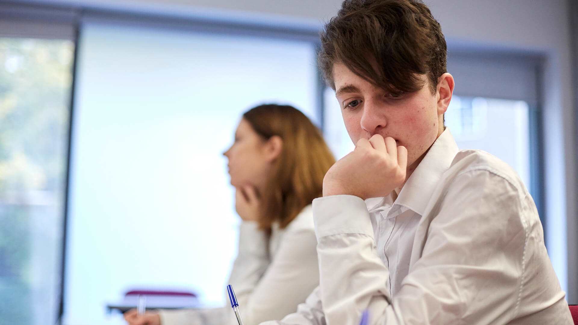 A student wearing uniform sits on a desk lost in a thought with their hand over their mouth, they sit next to another student who is focused on the lesson.