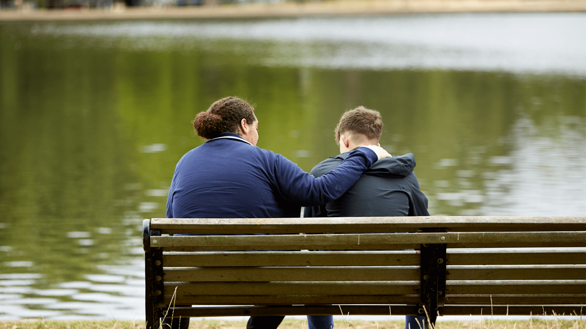 Two young people sitting on a bench in a park, one has their arm around the other.