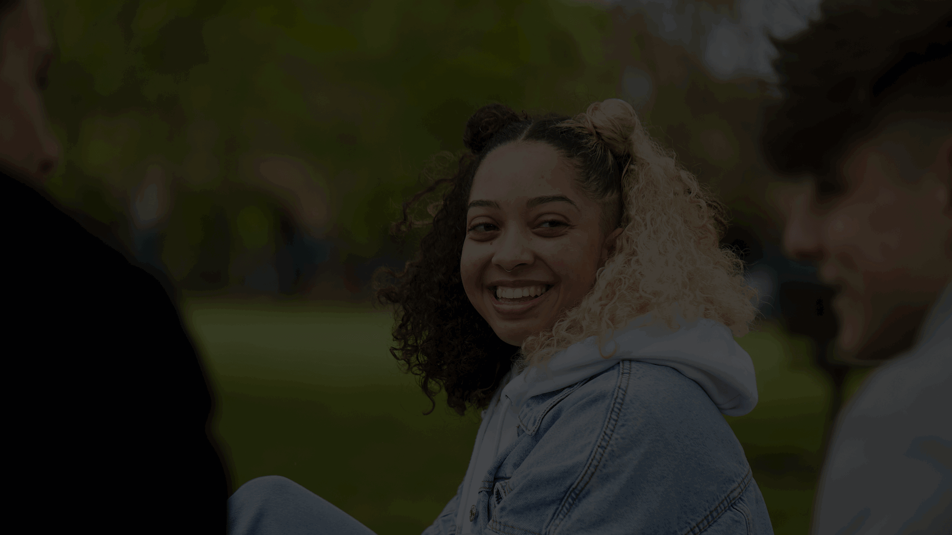 A young person is in the middle of the photo smiling while looking at another young person to the right. Another young person sits on the left looking at the young person in the middle. They are all sitting in a park with trees in the background.