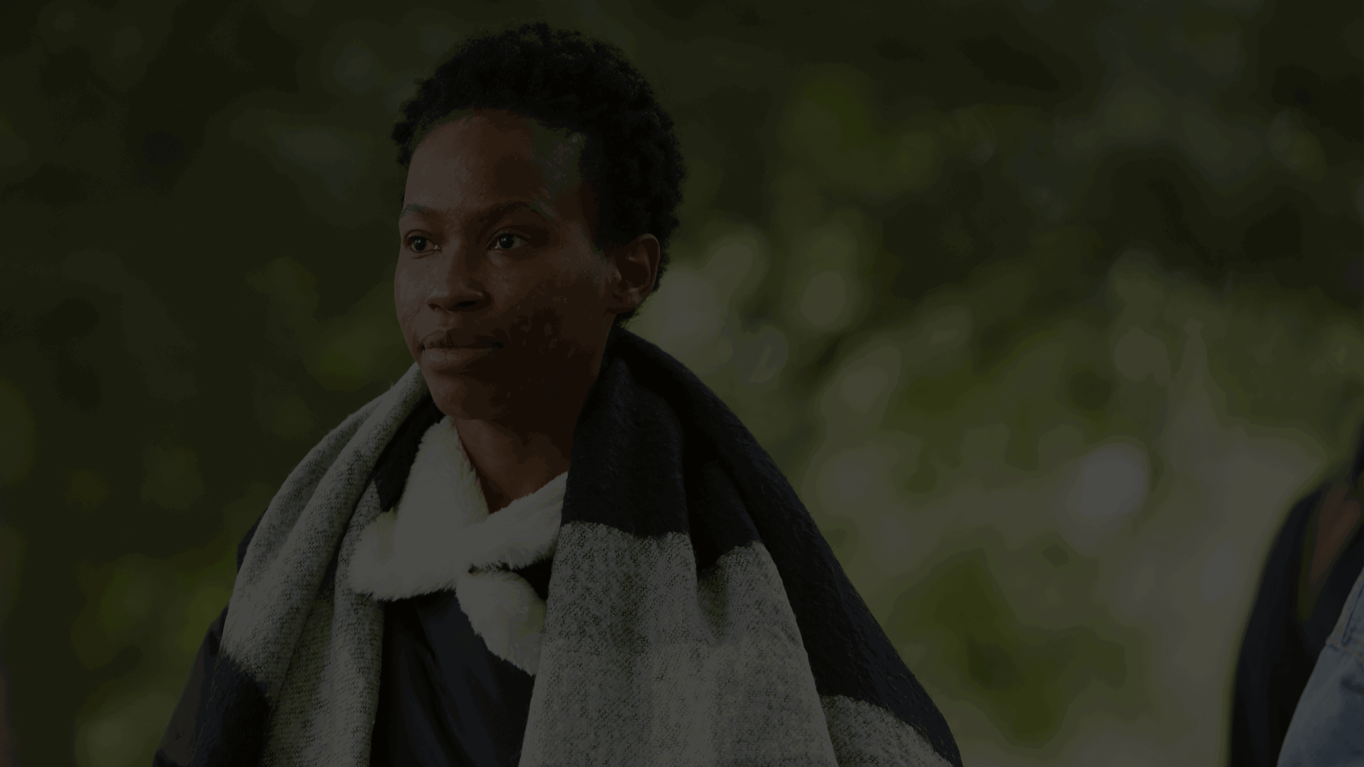 A young person walks through a park, looking forward with a neutral expression. They are wrapped up in a scarf and green trees sit in the background.