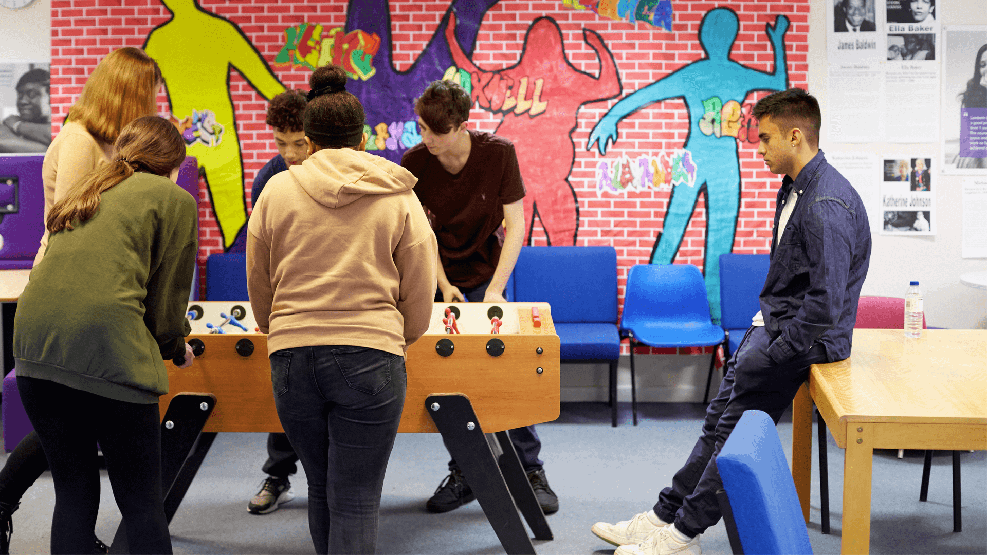 group-of-five-young-people-playing-table-football-while-one-boy-is-on-another-table-watching-with-graffiti-wall-on-background