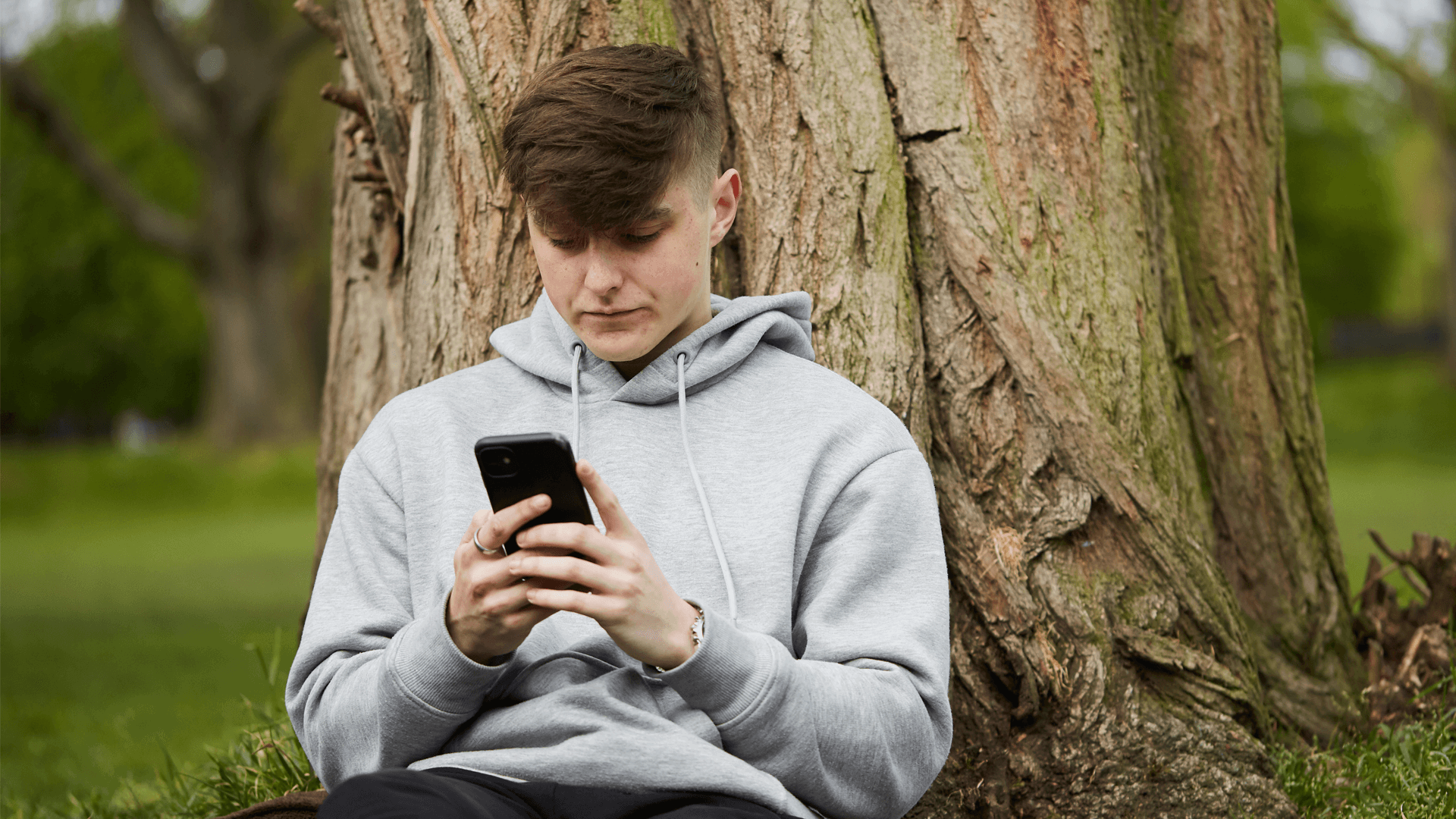 a boy wearing grey hoodie using his mobile phone while sitting on the ground leaning on a tree