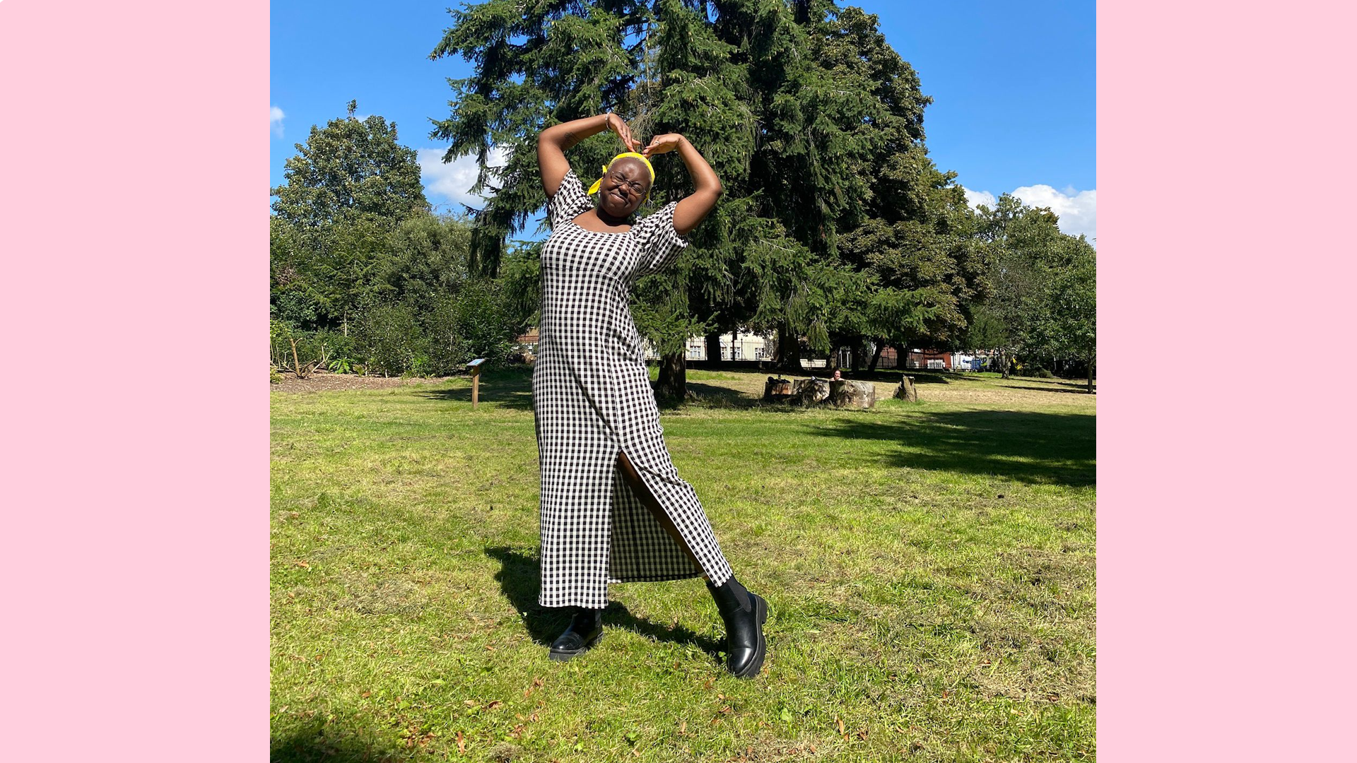 Jaleel is in a park in front of a big green tree. They have positioned their arms to create a heart shape.