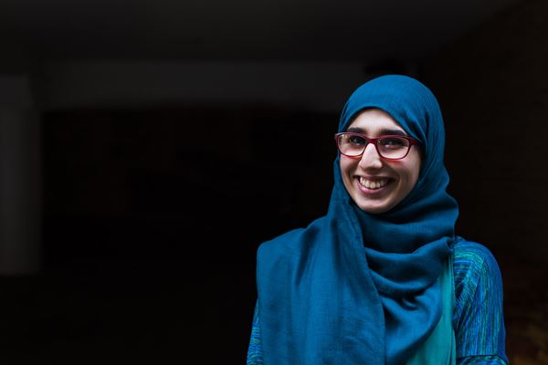 A young woman smiling and wearing a blue hijab and glasses.