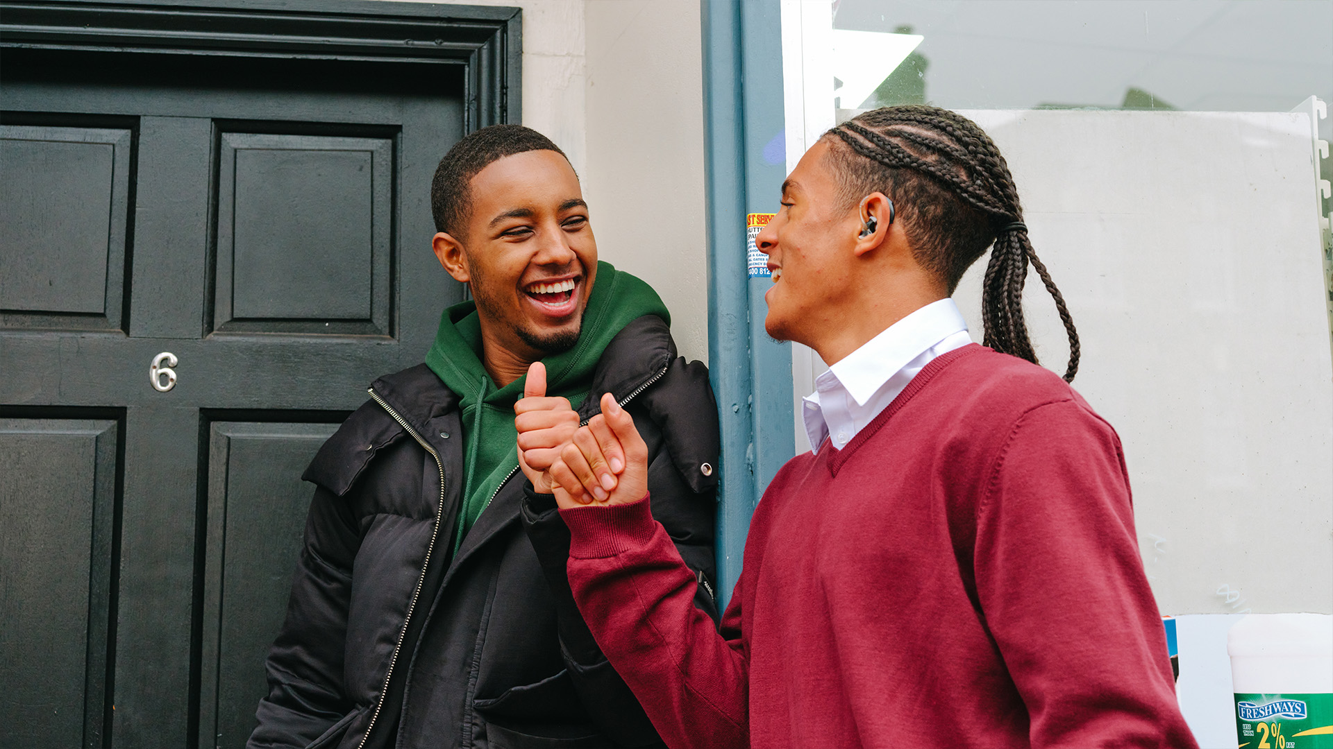 A Black teenage boy wearing a hearing aid bumping fists with a young Black man outside a front door.