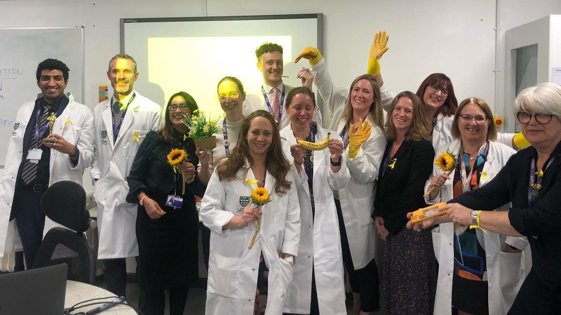 A group of teachers and staff from Weydon School, holding yellow flowers, bananas, and gloves.