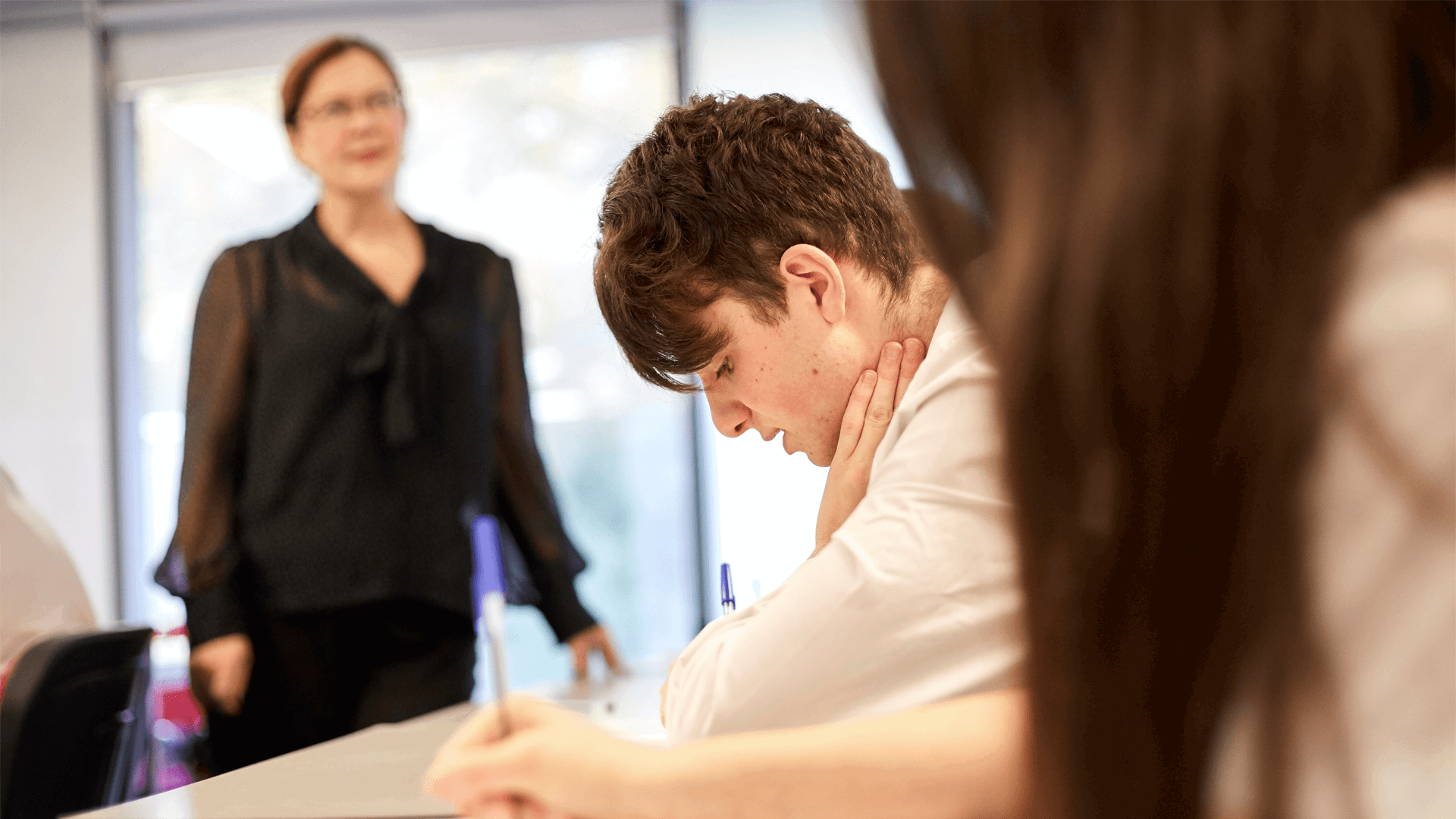 a-boy-in-a-uniform-sitting-on-his-desk-writing-on-his-desk-and-hands-on-his-neck-while-exams-is-on-going-in-class-a-teacher-is-standing-on-the-background