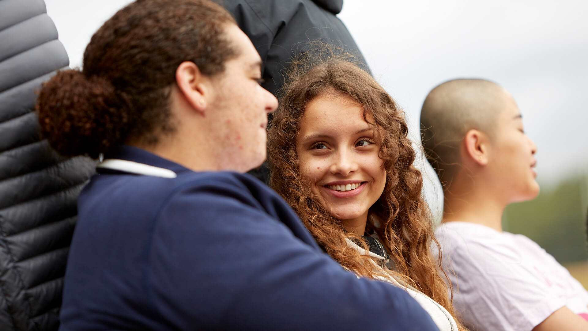 Three young people sit on a bench outside. The person on the right is chatting with the person in the middle of the bench who is looking at them and smiling. The person on the right is speaking to someone outside of the image.