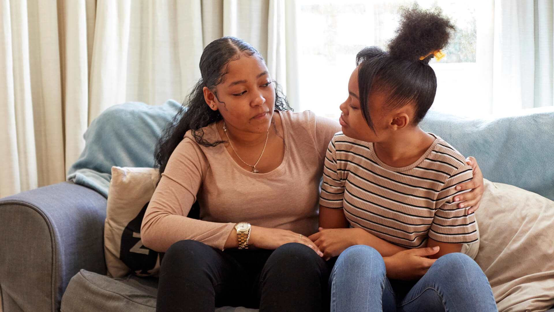 Two young people sit on a sofa with the person on the left putting an arm around the other. They both are looking at each other while talking.