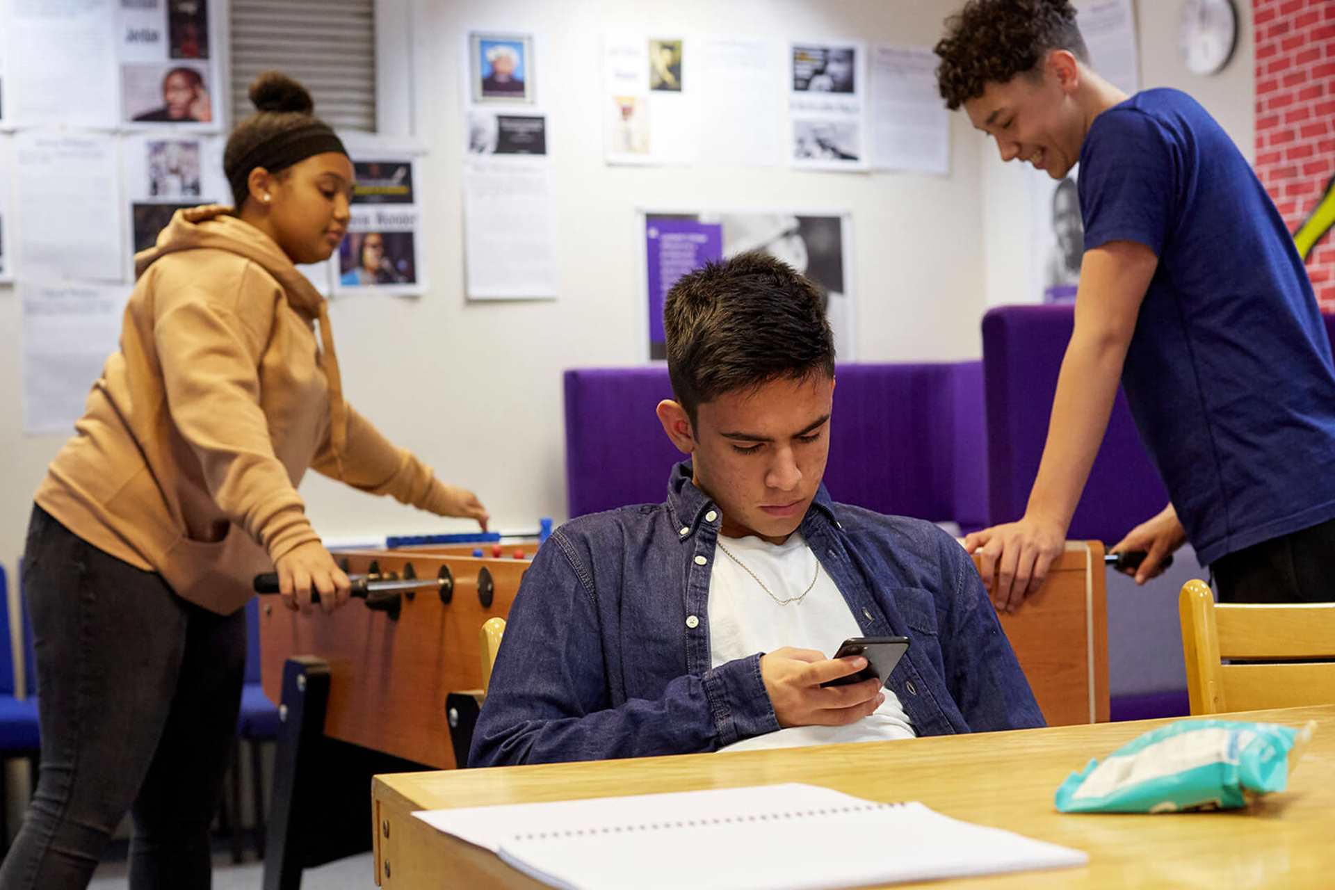 A young person is looking seriously down at his phone at a table in a common room. There are two young people behind him playing table top football, one of them is laughing.