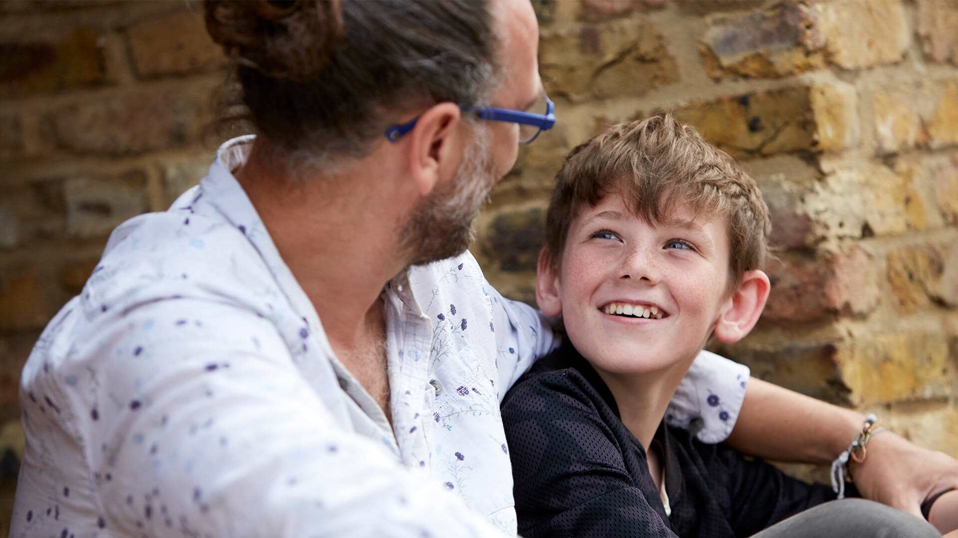 Father and son smiling and looking very happy together sitting by a wall