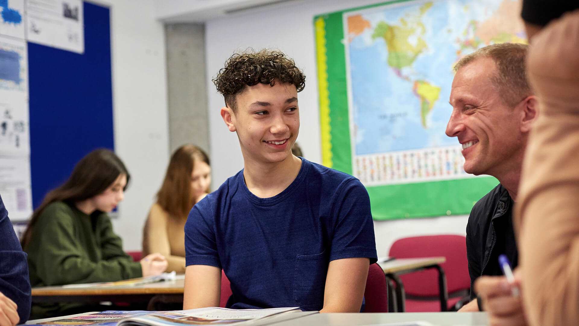 a student sitting on his desk smiles on his teacher who is sitting behind him to encourage him on school work