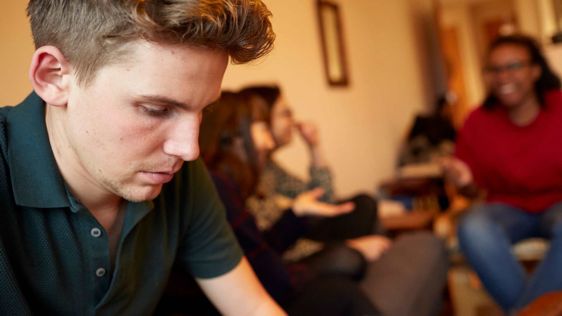 medium-shot-of-a-young-man-looking-down-feeling-alone-while-group-of-friends-are-talking-and-laughing-on-the-background