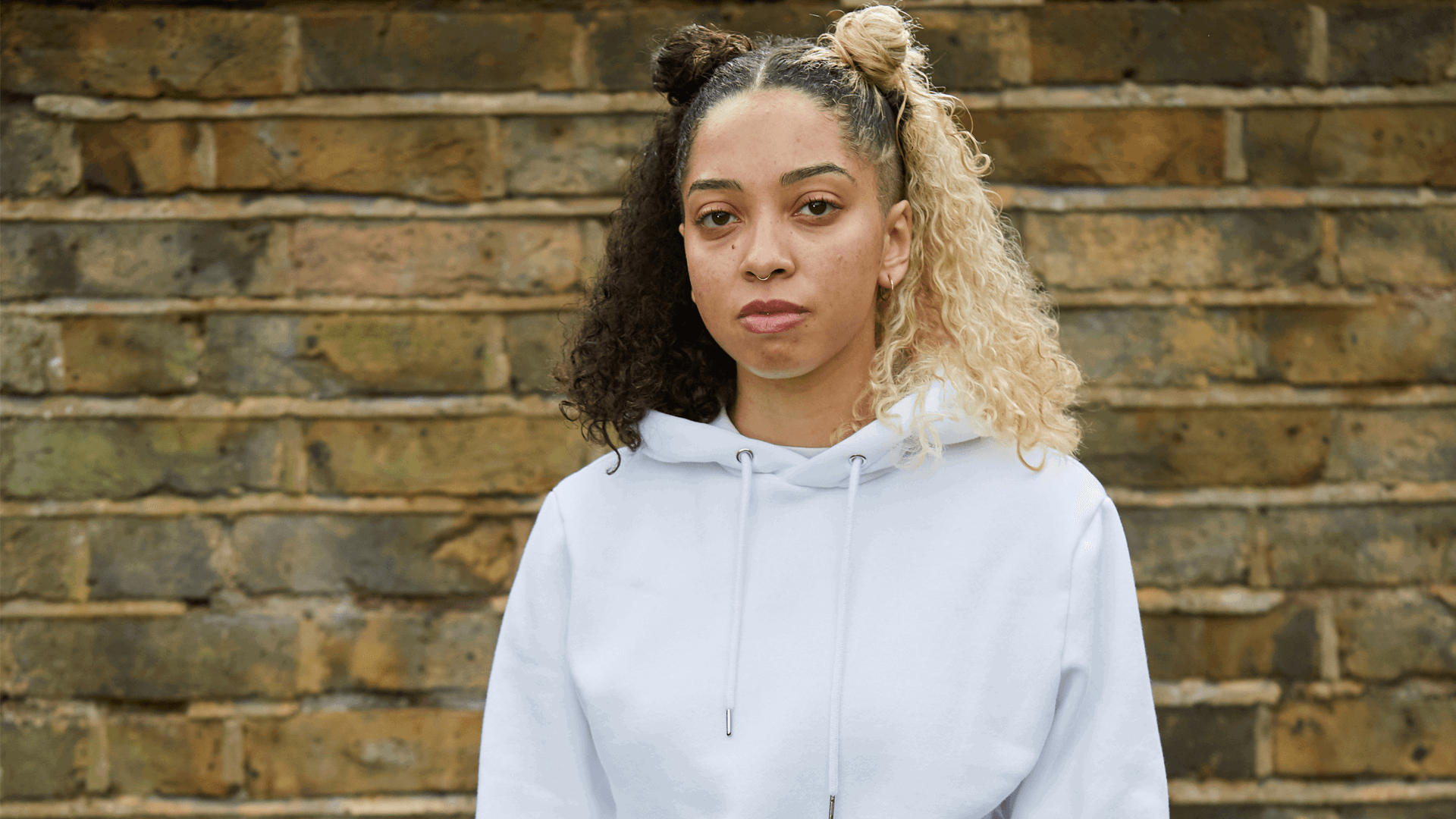 medium shot of a girl with curly hair wearing white hoodie looking in front of the camera with brick wall on the background