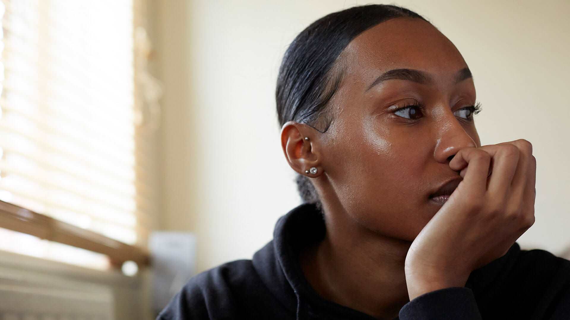 A young person sits in a room wearing a black hoodie and their hair tied back. They are looking to the right with their hand curled over their mouth, lost in thought.