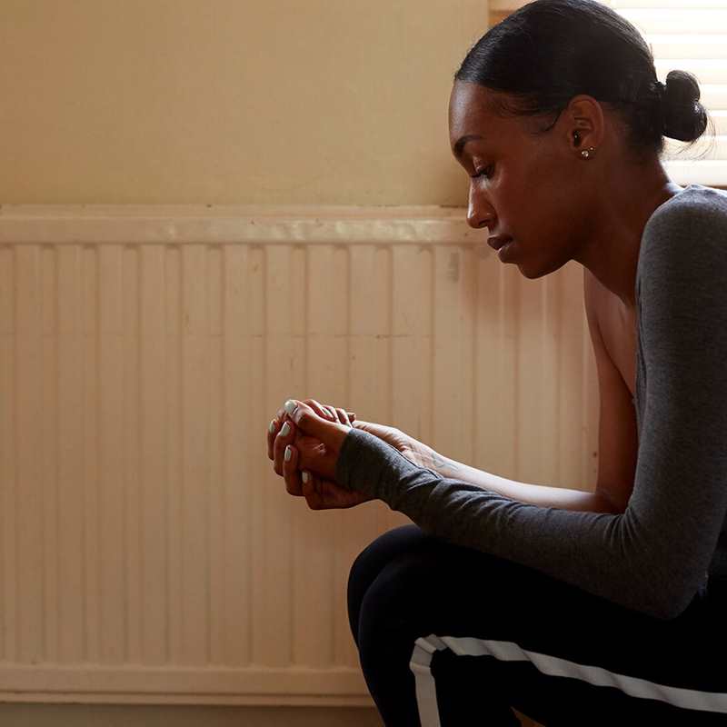 medium-shot-of-a-girl-sitting-in-a-room-beside-a-radiator-while-thinking-and-looking-at-her-hands-clasped-together