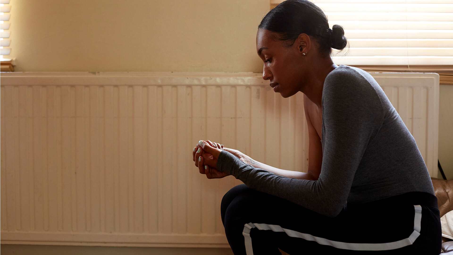 medium-shot-of-a-girl-sitting-in-a-room-beside-a-radiator-while-thinking-and-looking-at-her-hands-clasped-together