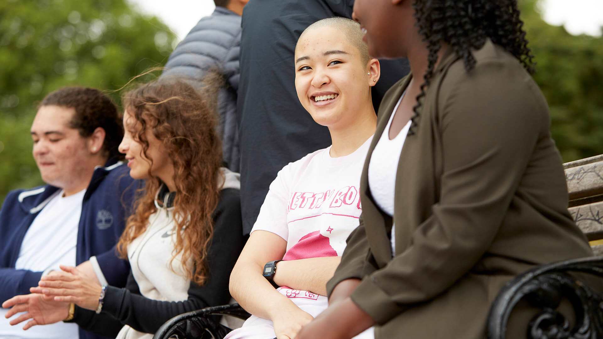 A group of young people sit on a bench together. Two of them are sitting on top of the bench while the other four sit on the seat of the bench. They are in pairs, talking to one another. The focus of the image is the person in the middle who wears a pink t-shirt and is smiling at the person next to them on the right.