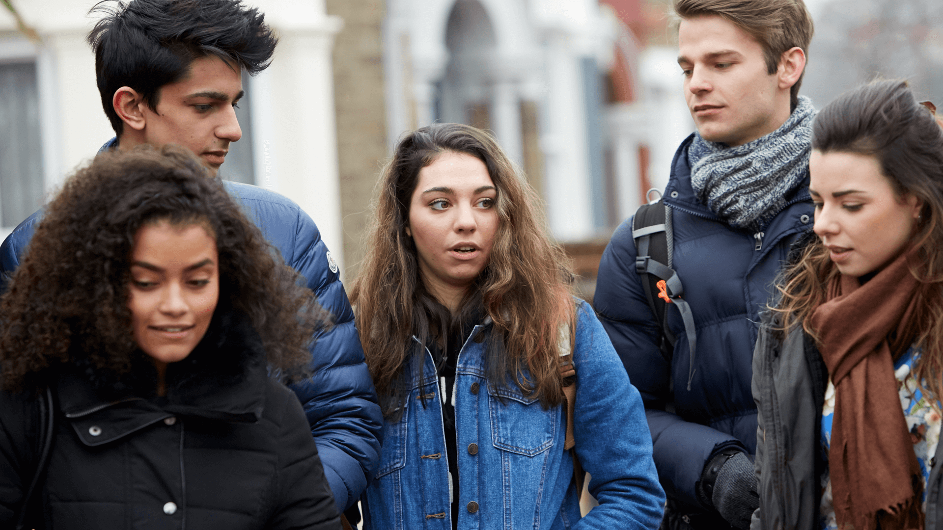 Five young people wearing coats, jackets and backpacks. They are talking as they walk down a street.