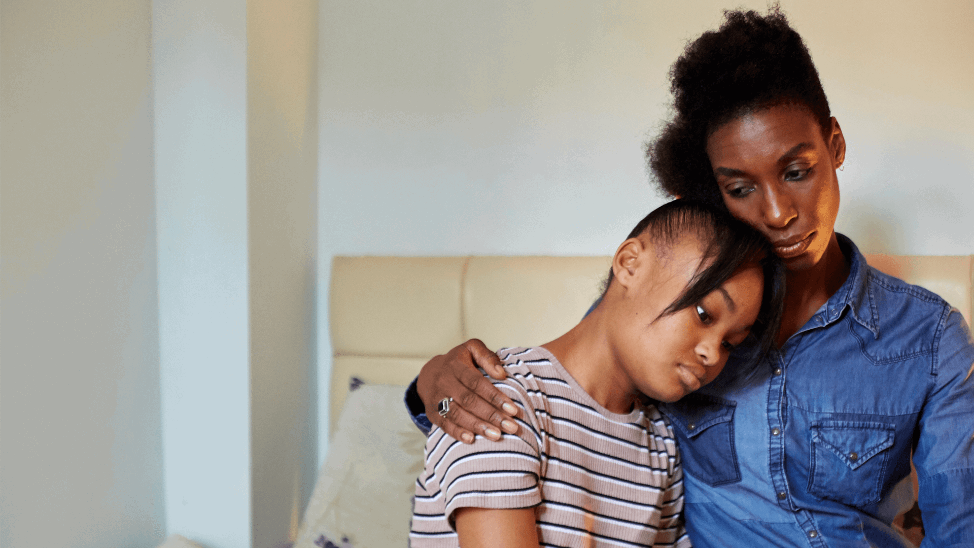 A mother putting her arm around her sad daughter in a bedroom