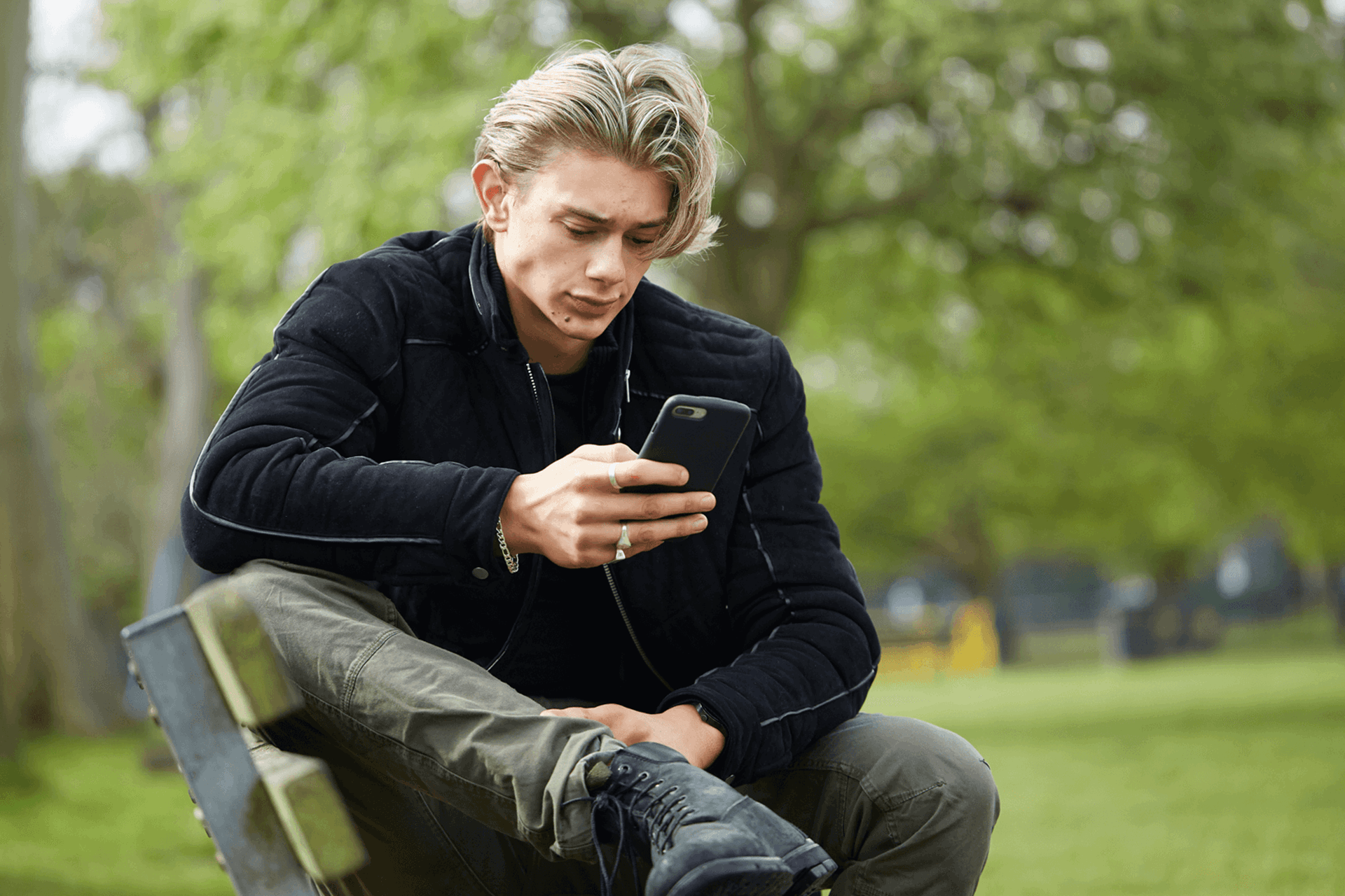medium-shot-of-a-young-man-in-black-jacket-looking-at-his-phone-while-sitting-on-a-bench-in-the-park-with-trees-on-background