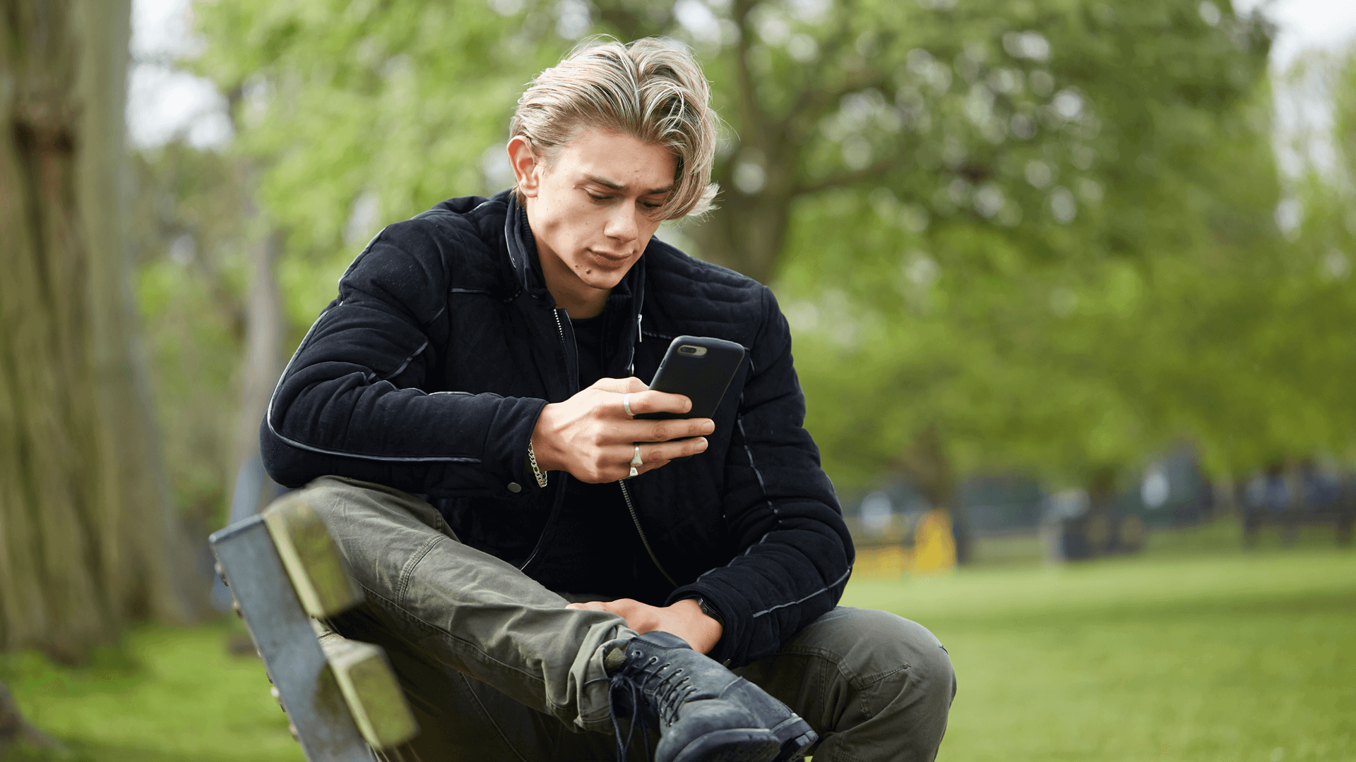 medium-shot-of-a-young-man-in-black-jacket-looking-at-his-phone-while-sitting-on-a-bench-in-the-park-with-trees-on-background