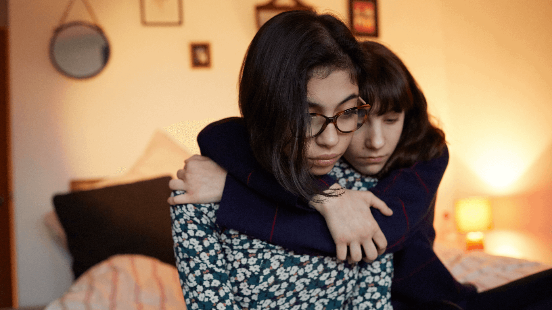 medium shot of a girl hugging another girl who is upset in the bedroom with night light on and hanging photo frames on background