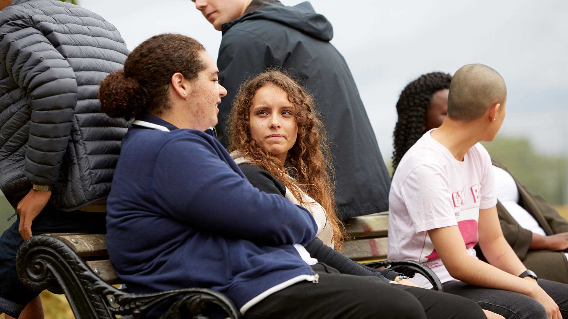 group-of-six-young-people-sitting-on-bench-talking-with-focus-on-a-girl-and-a-boy-talking-to-each-other