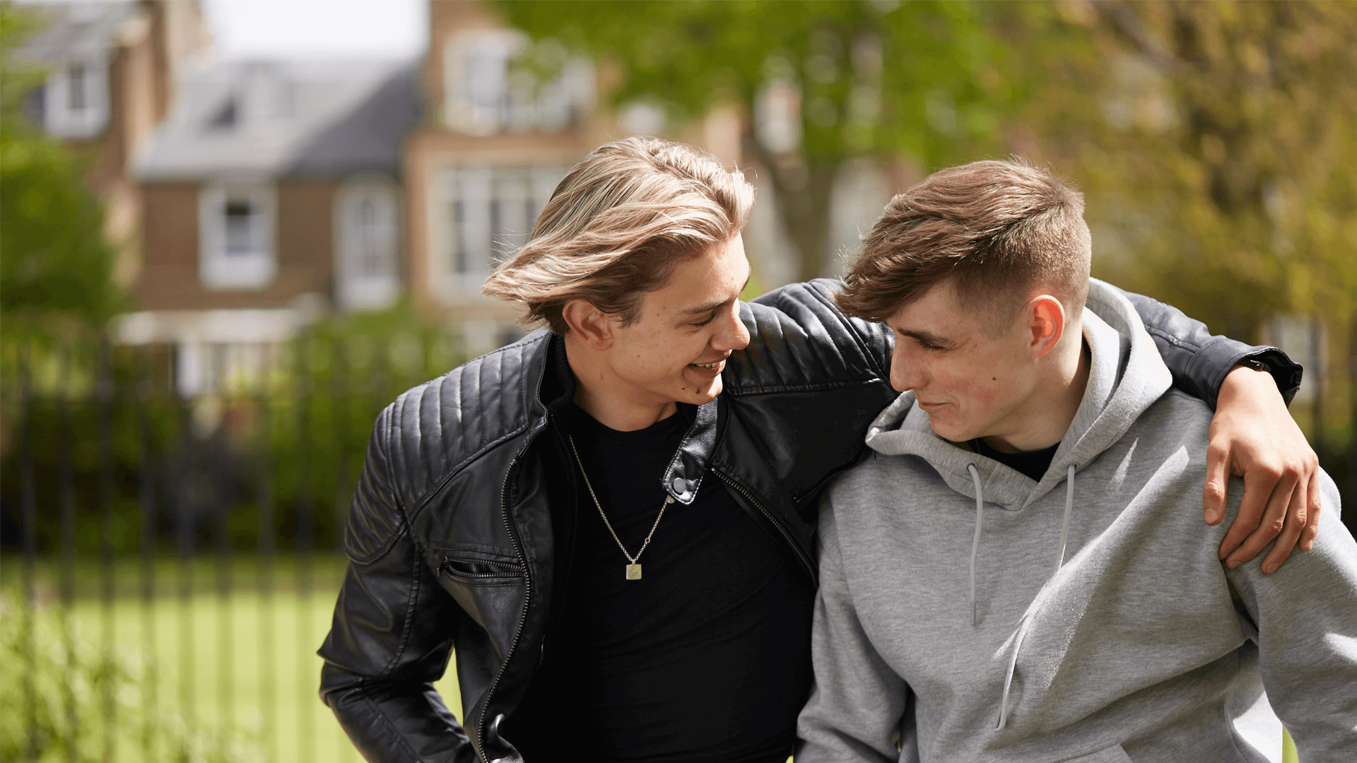 a boy wearing black jacket smiling while wrapping his arm to his friend wearing grey jacket smiling back to him with houses and trees on background