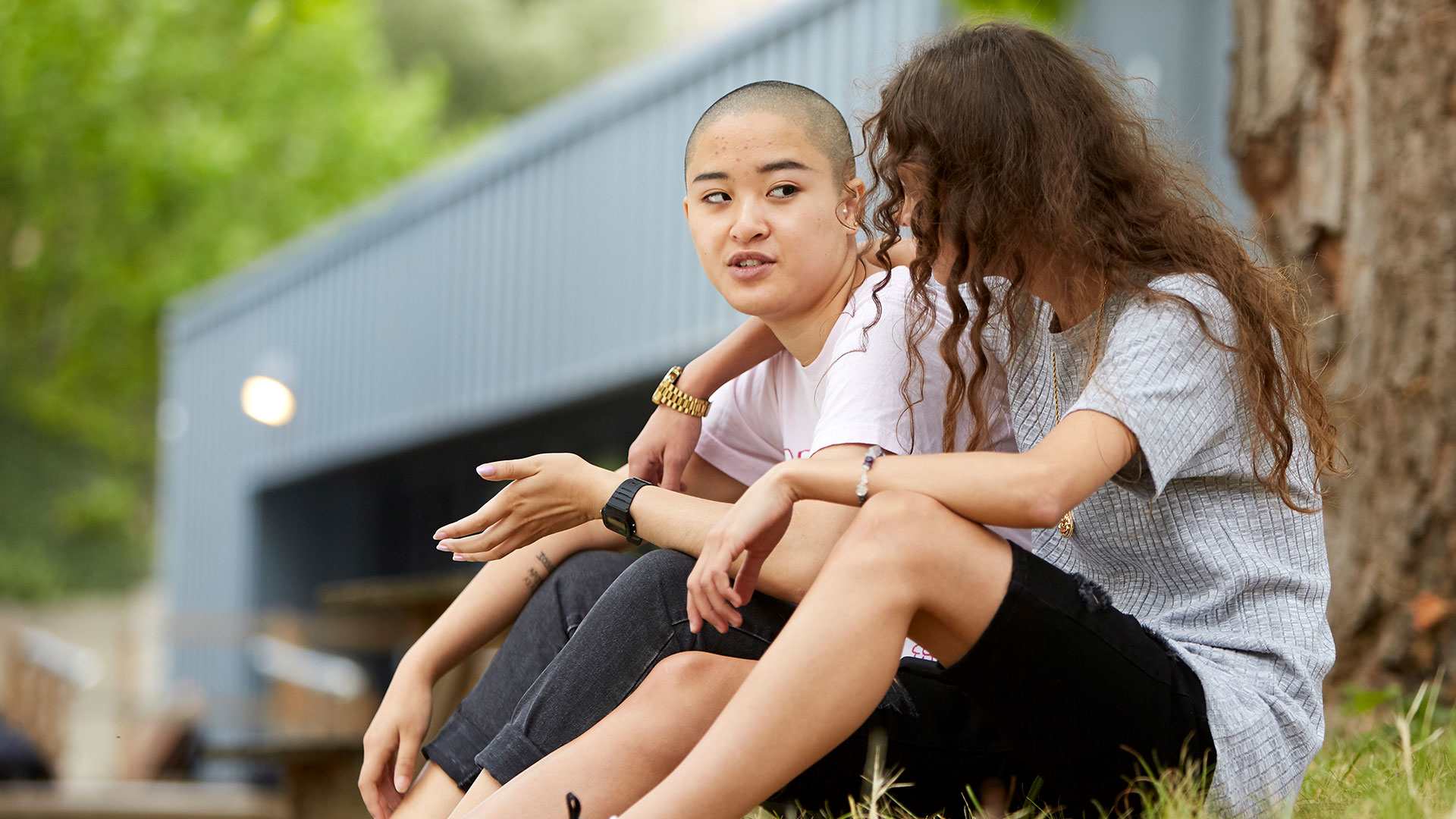 a-young-woman-with-curly-hair-and-in-grey-shirt-and-black-shorts-with-face-unseen-has-her-hands-wrapped-another-young-woman-with-shaved-hair-and-in-white-shirt-and-blakc-trousers-talking-to-her-while-they-sat-on-the-grass-with-a-tree-and-building-on-their-background