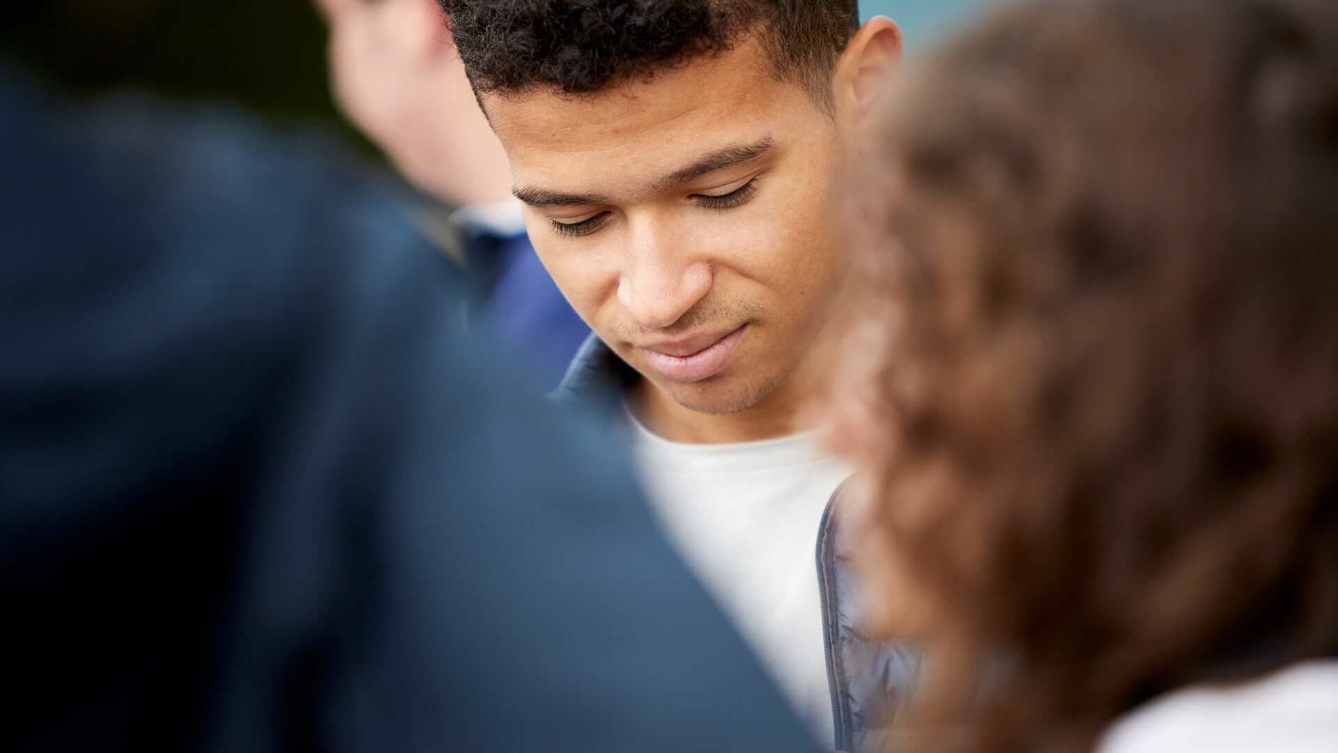 close-up-of-a-young-man-in-white-shirt-and-blue-jacket-looking-down-with-eyes-closed-and-a-girl-with-curly-hair-and-another-person-in-the-foreground