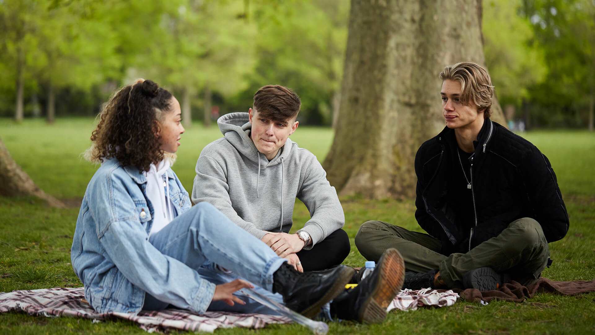 wide-shot-of-three-young-people-sitting-on-the-ground-together-talking-to-each-other-and-listening-with-trees-on-their-background