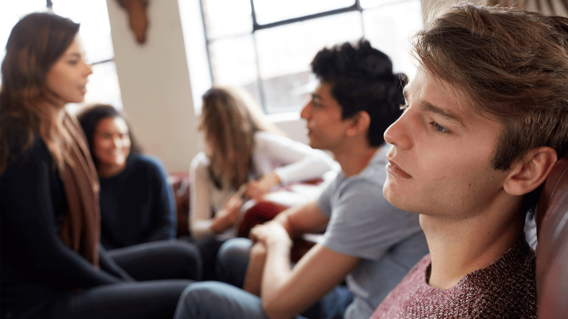 a boy lost in thoughts while sitting on the couch with friends sitting beside him talking to each other