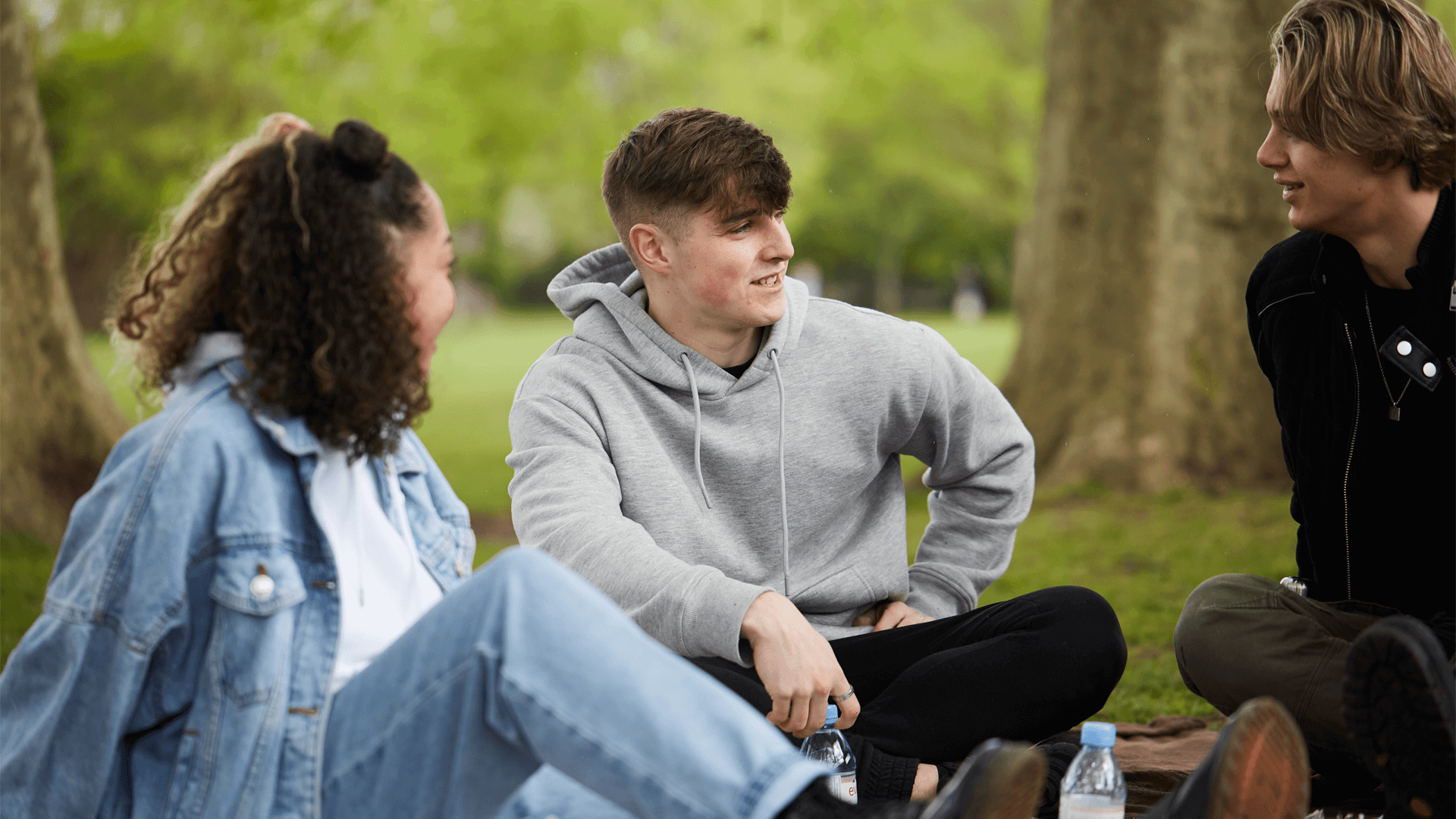 a-group-of-three-young-people-talking-at-each-other-while-sitting-on-the-ground-with-trees-of-the-park-on-background