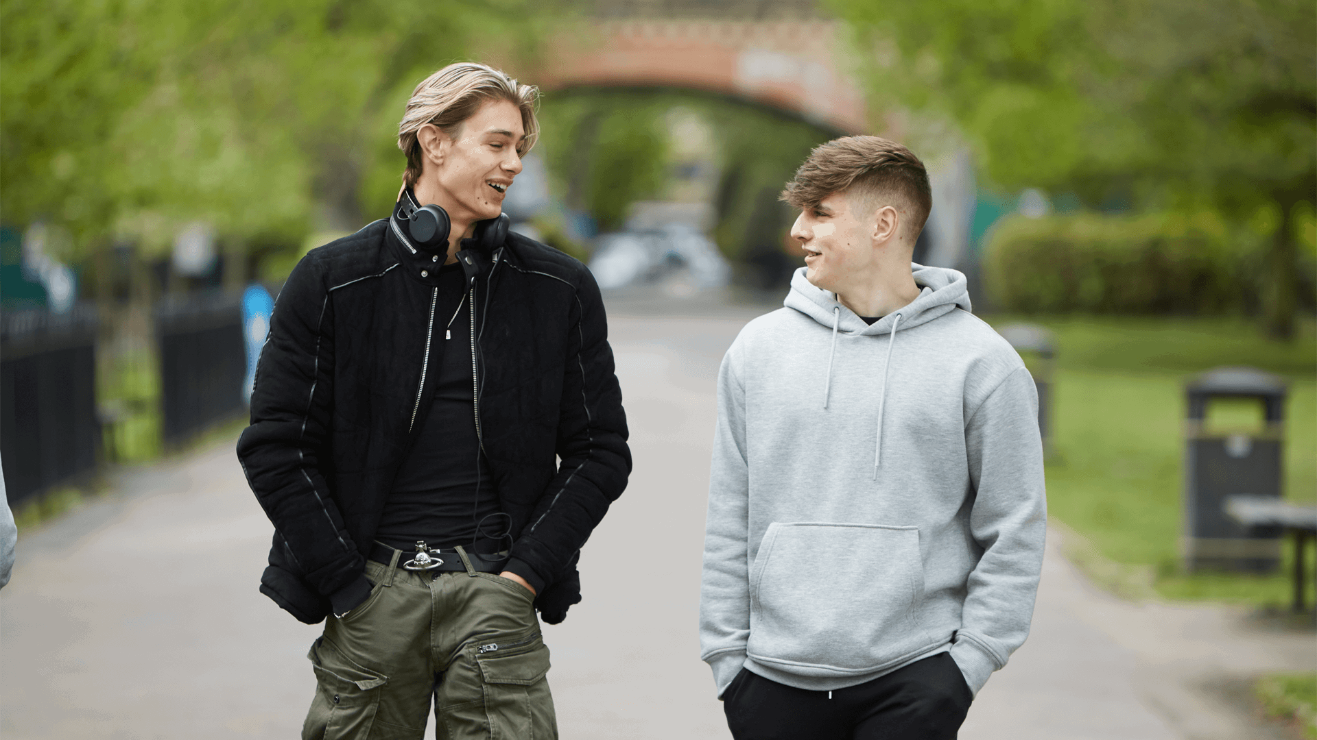 two-young-man-wearing-black-jacket-with-headphones-on-his-neck-and-hands-on-the-pockets-of-his-trousers-talking-while-walking-with-another-boy-wearin-grey-hoodie-on-a-street
