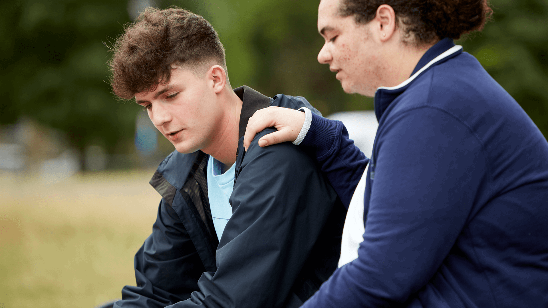 a boy comforting his upset friend putting with his hand on his shoulder while they sit in a park