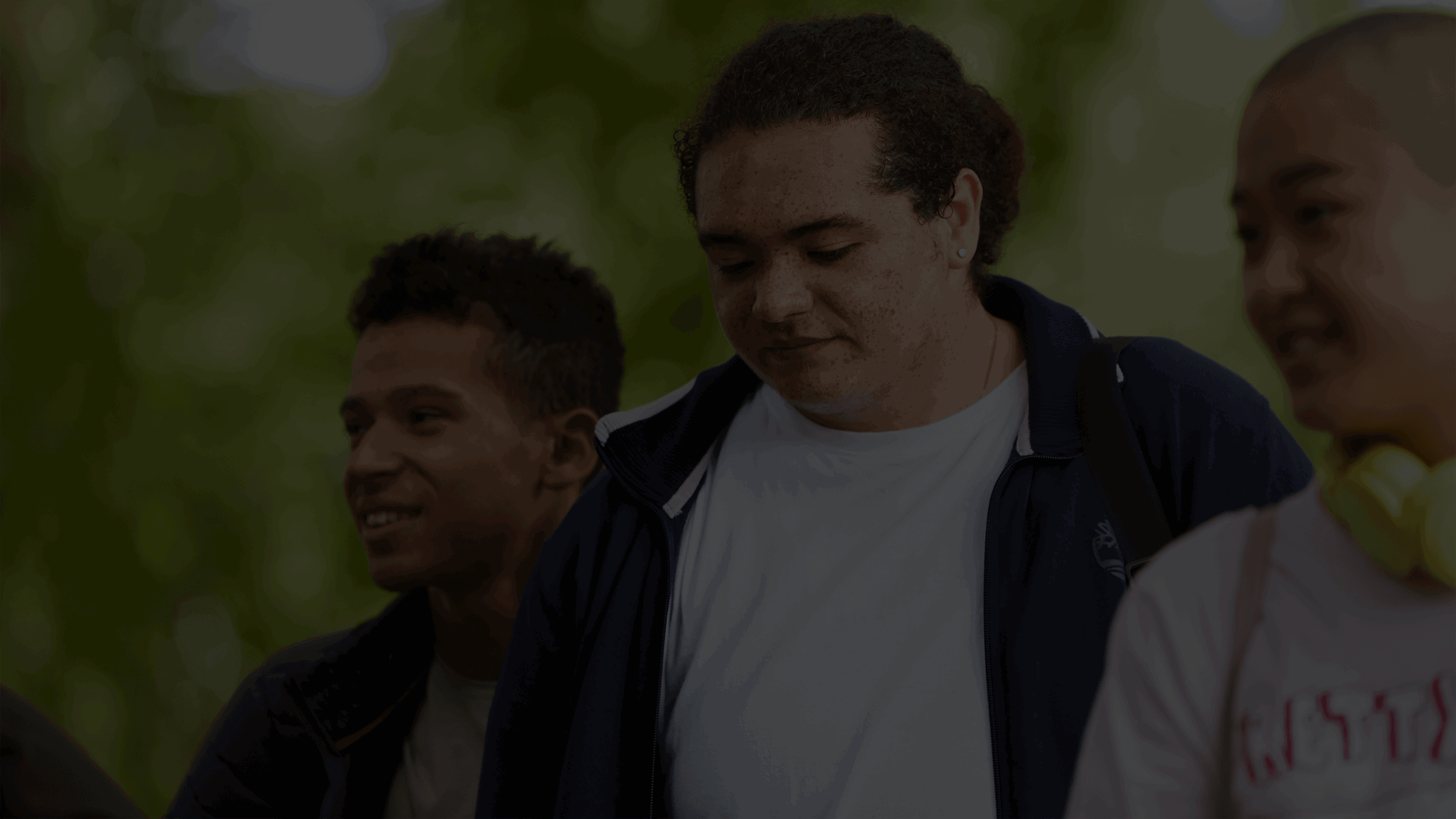 Three young people walk together through a park. On the left the young person is at the back looking to the right. The young person in the middle is looking down. The young person at the front on the right also looks to the right and is wearing yellow headphones.