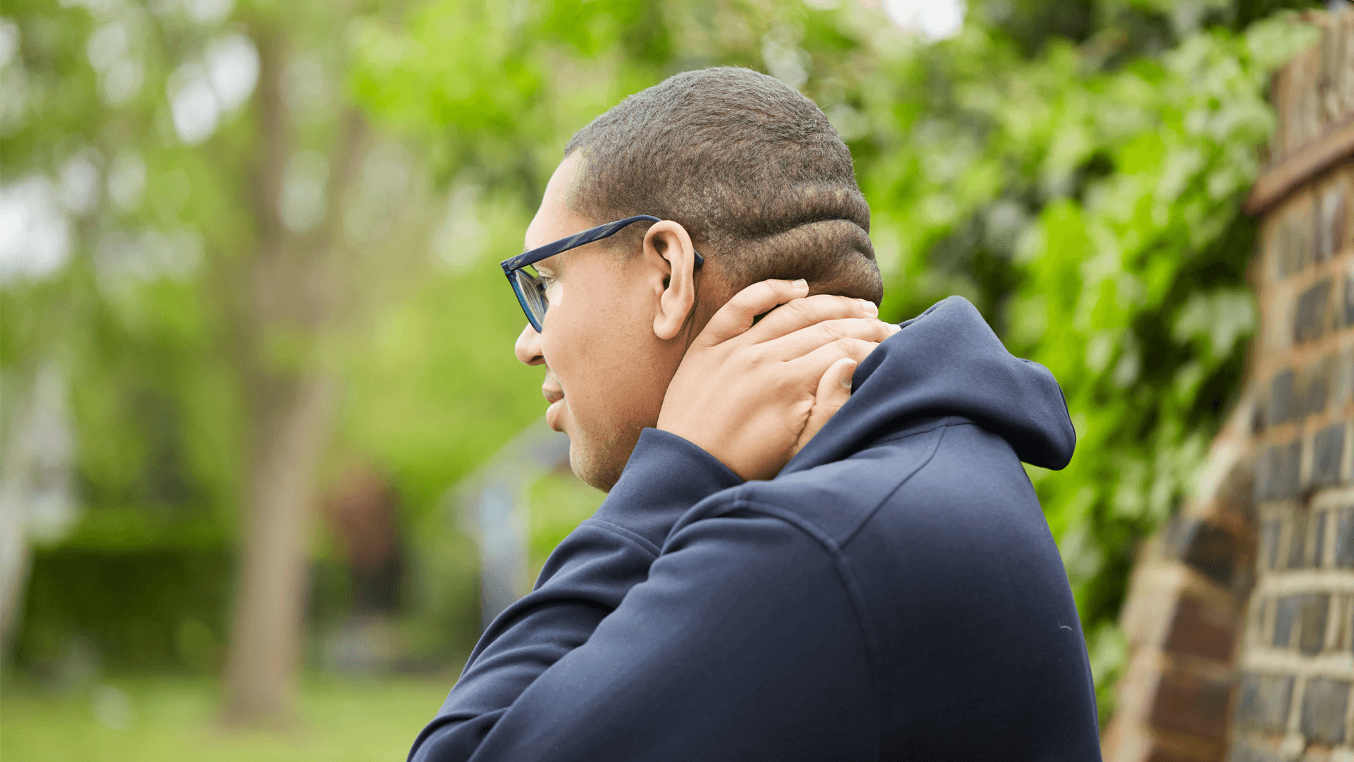 side view close up of a boy wearing glasses and black hoodie looking worried as one of his hands hold his neck