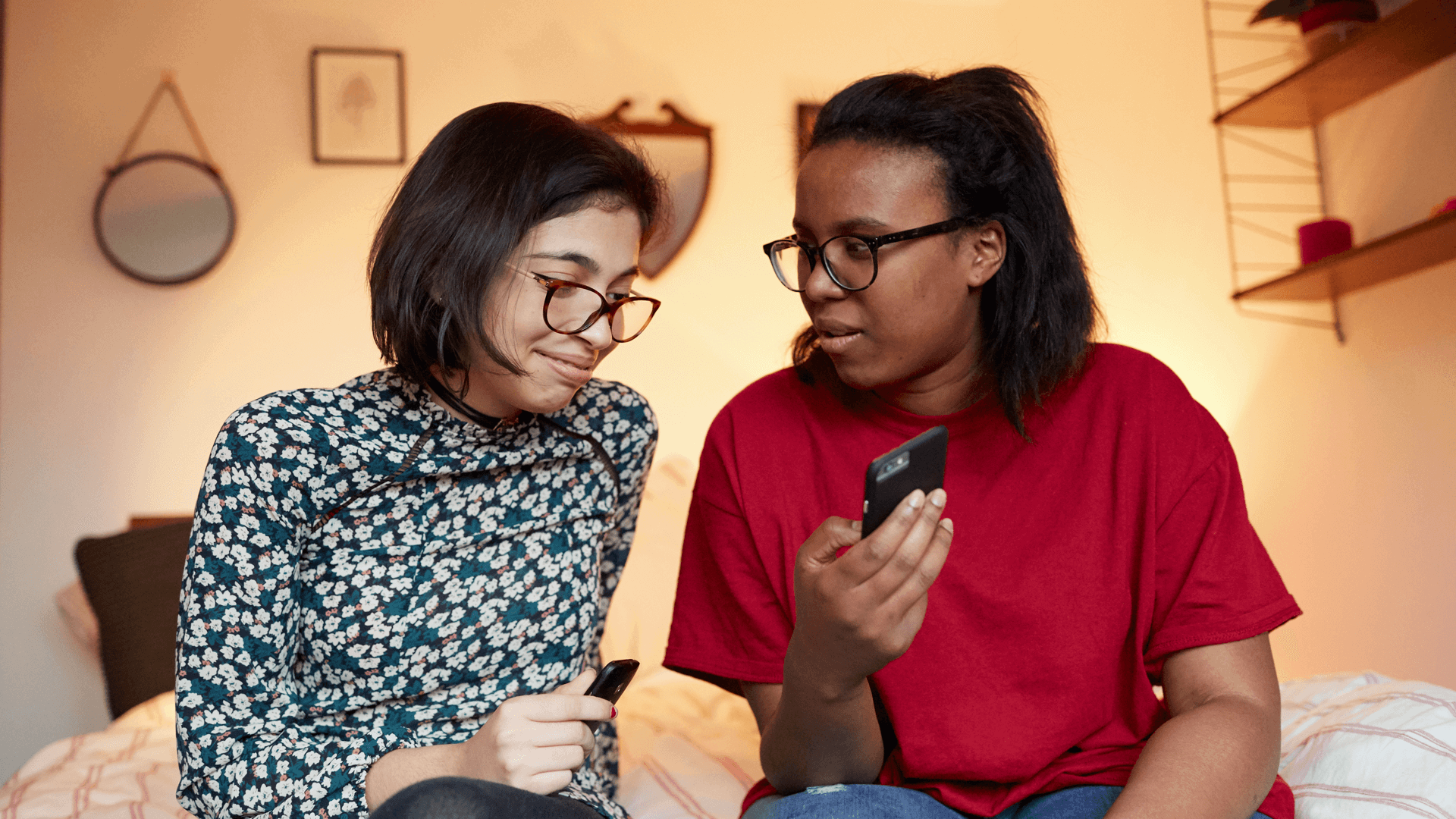 two-girls-sitting-in-a-bed-one-girl-is-showing-her-phone-to-another-girl-while-she-reacts-on-what-she-sees-on-the-phone